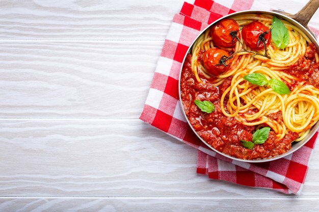 Primer plano de espaguetis de pasta con carne y salsa boloñesa de tomate en una sartén servida con albahaca y tomates cherry sobre fondo rústico de madera blanca, vista superior. Cena tradicional italiana, espacio para texto