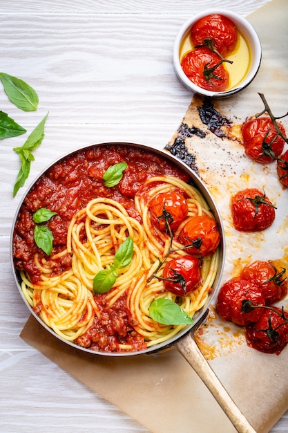 Primer plano de espaguetis de pasta con carne y salsa boloñesa de tomate en una sartén servida con albahaca y tomates cherry sobre fondo rústico de madera blanca, vista superior. Cena tradicional italiana