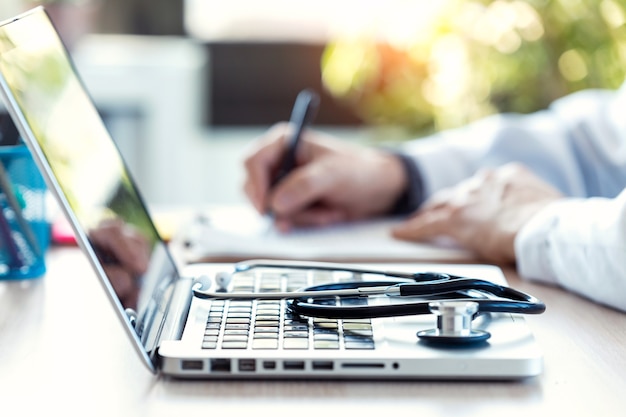 Primer plano de un estetoscopio médico en el teclado de la computadora portátil. Desenfoque de fondo de un médico que revisa la documentación de un paciente en consulta.