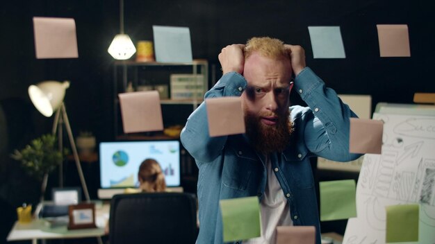 Foto primer plano estresado tipo hipster agarrando la cabeza en la oficina de noche empresario exhausto poniéndose nervioso por el proyecto en la oficina de hipster empleado sorprendido mirando pegatinas en el tablero de vidrio en el interior