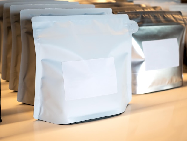 Primer plano etiqueta blanca en blanco vacía en bolsas modernas de papel de aluminio blanco y plateado empaque de granos de café Etiqueta adhesiva de plantilla de maqueta Fila de empaque de bolsa con cremallera de papel para granos de café que se muestran en el estante