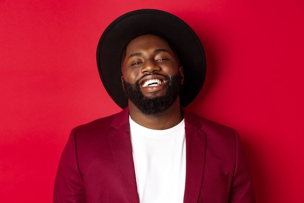 Primer plano de feliz guapo negro sonriéndole, mirando complacido, con sombrero negro y chaqueta, fondo rojo.