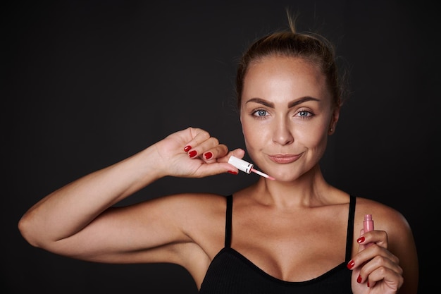 Primer plano foto de estudio de mujer rubia de mediana edad adorable encantadora encantadora femenina con piel bronceada fresca sana y tubo natural de brillo de labios rosa Concepto de corrección de labios de maquillaje mimos