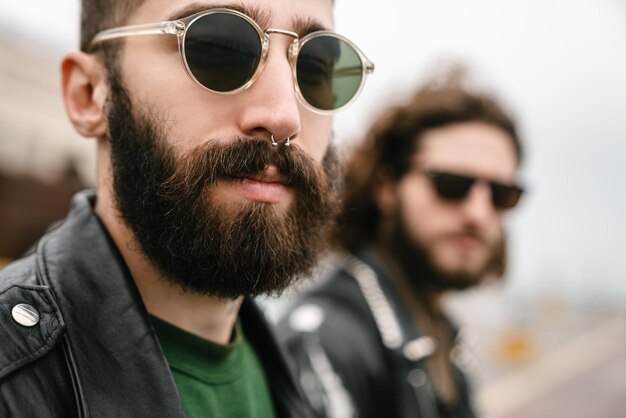 Primer plano de la foto de los hombres caucásicos barbudos ciclistas en gafas de sol posando y mirando a la cámara juntos al aire libre