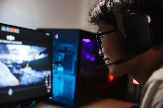 Primer plano de la foto de un joven jugador adolescente alegre jugando videojuegos en línea en la computadora en una habitación oscura, usando auriculares con micrófono y usando un teclado colorido retroiluminado