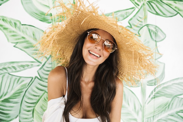 Primer plano de la foto de una mujer bonita morena con sombrero de paja y gafas de sol sonriendo a la cámara mientras está de pie sobre la pared colorida