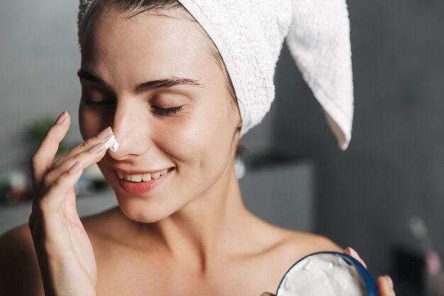 Primer plano de la foto de la mujer complacida envuelta en una toalla en la cabeza aplicando crema en la cara