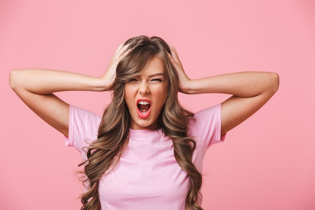 Primer plano de la foto de una mujer enojada molesta con el pelo largo y rizado en una camiseta básica que cubre las orejas y gritando debido al ruido terrible, aislado sobre fondo rosa