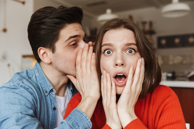 Primer plano de la foto de una mujer joven que mantiene las manos en la cara con sorpresa, mientras que el hombre le susurra un secreto al oído durante la cita en el café