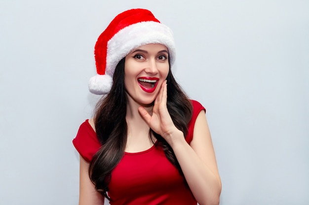 Primer plano de la foto de Navidad de una chica impresionada con Wow gritando OMG en vestido rojo y sombrero de Santa en el espacio gris.