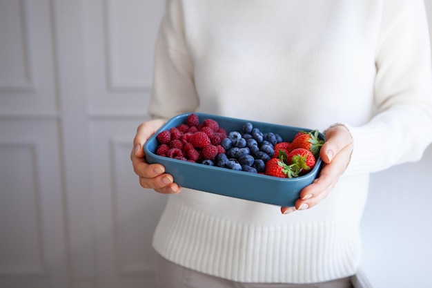 Primer plano de frambuesas, fresas, arándanos en un tazón azul en manos de una mujer con suéter blanco en un fondo claro. Copie el espacio y la profundidad de campo