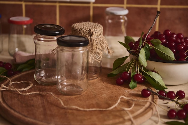 Primer plano de frascos estériles con tapas en tablero de madera junto a un recipiente con cerezas recién recogidas en la encimera de la cocina