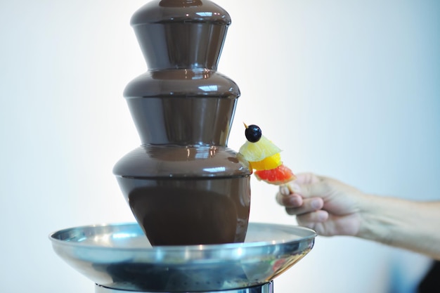 Primer plano de una fuente de chocolate líquido y frutas frescas en un palo sostenido a mano en el evento de la fiesta de bodas