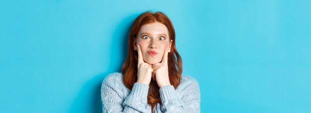 Primer plano de una graciosa jovencita pelirroja haciendo muecas entrecerrando los ojos y picando las mejillas de pie contra el azul b