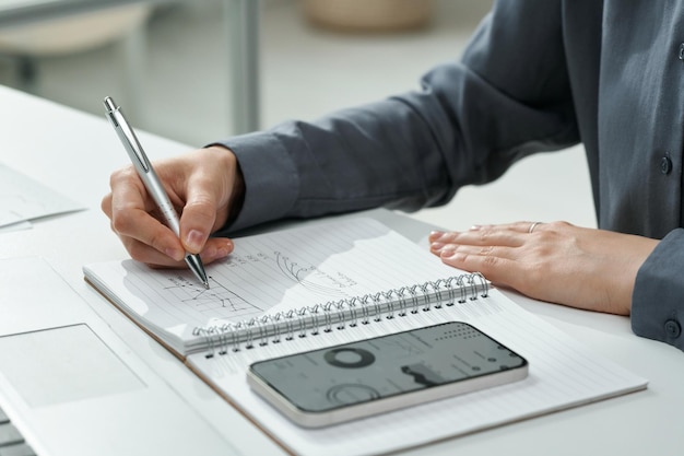 Primer plano de gráficos de dibujo financiero en el bloc de notas usando su teléfono inteligente mientras trabaja en la mesa en la oficina