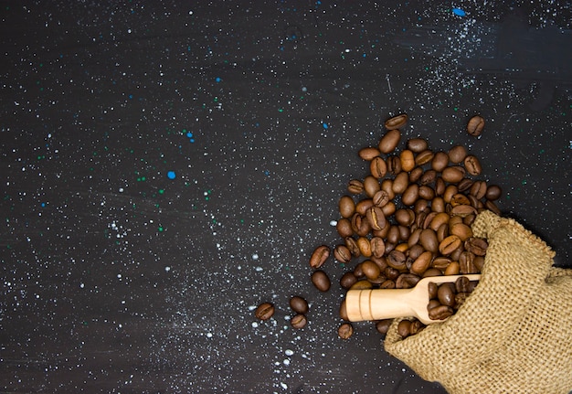 Primer plano de granos de café tostados vertidos en una bolsa de arpillera sobre un fondo oscuro, vista superior. Pala de madera al lado de la bolsa.