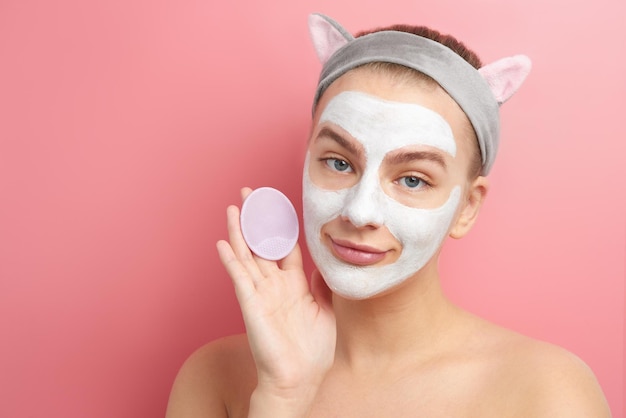 Primer plano de una hermosa joven con máscara blanca y diadema de orejas de gato coreano sonriendo y sostiene un cepillo facial, cuidado de la piel en estudio rosa