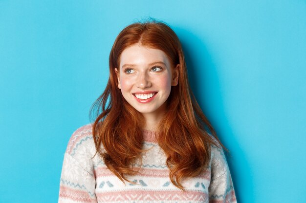 Primer plano de la hermosa modelo de mujer pelirroja en suéter de invierno, sonriendo y mirando a la izquierda en el espacio de la copia, de pie sobre fondo azul