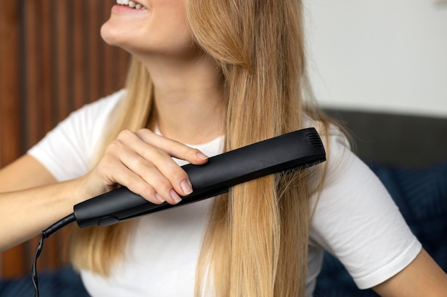 Primer plano de una hermosa mujer sonriente usando plancha de pelo en casa enfoque selectivo en el cabello