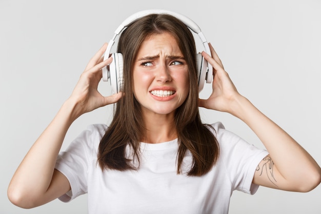 Foto primer plano de una hermosa niña molesta en auriculares inalámbricos quejándose de música horrible, haciendo muecas molestas.