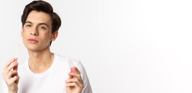 Foto primer plano de una hermosa persona queer con brillo en la cara aplicando brillo de labios y mirando seguro a la cámara de pie sobre fondo blanco