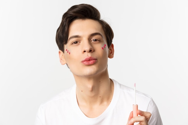 Primer plano de un hermoso hombre gay con brillo en los labios fruncidos de la cara después de aplicar brillo de labios, de pie sobre blanco.