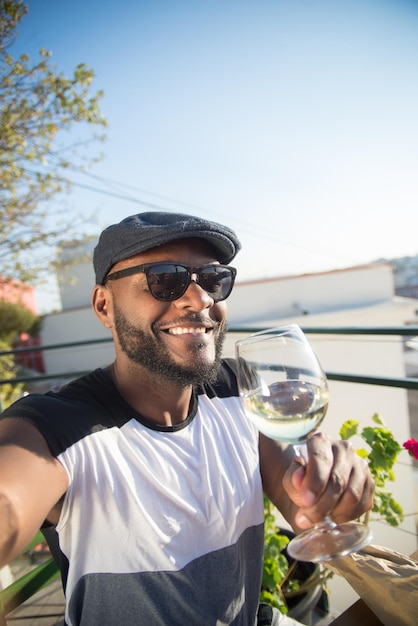 Primer plano de un hombre africano feliz sonriendo para la cámara del teléfono. Joven barbudo con gorra negra sosteniendo una copa de vino posando y mirando la cámara del teléfono inteligente. Momentos felices y concepto de tecnologías modernas.