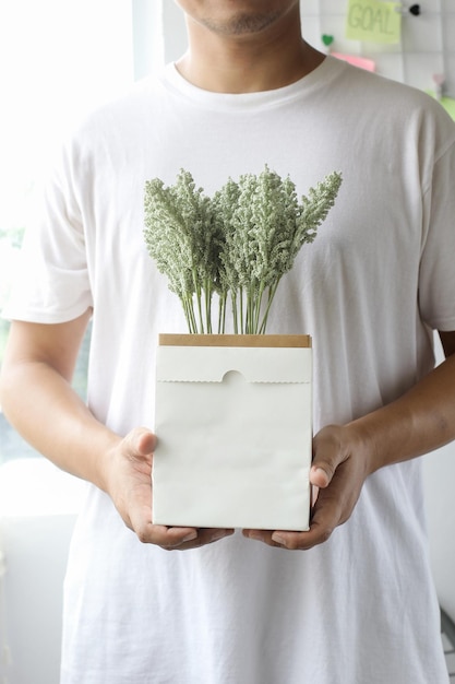 Primer plano de un hombre asiático con camiseta blanca sosteniendo un ramo de flores en un jarrón de bolsa de papel blanco en blanco para moc