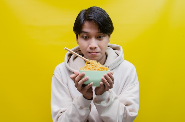 Primer plano, un hombre asiático con una capucha beige come fideos instantáneos con palillos y un tazón