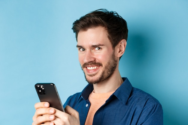 Primer plano de hombre barbudo alegre con smartphone, sonriendo confiado a la cámara, de pie sobre fondo azul. Copia espacio