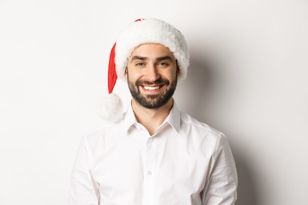 Primer plano de hombre barbudo feliz celebrando la Navidad, vistiendo gorro de fiesta de santa y sonriendo, de pie sobre fondo blanco.