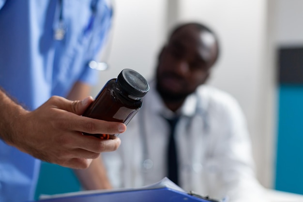 Primer plano de un hombre caucásico enfermera sosteniendo la botella de píldoras en la mano analizando la prescripción de medicamentos trabajando en la oficina del hospital. Médico afroamericano explicando el tratamiento médico. Servicios de salud