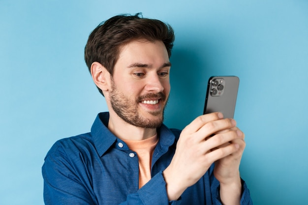 Primer plano de un hombre caucásico guapo escribiendo un mensaje, leyendo la pantalla del móvil y sonriendo, de pie sobre fondo azul.