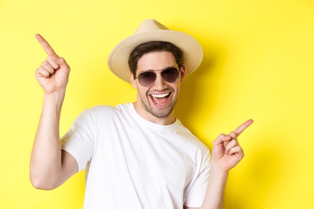 Primer plano del hombre disfrutando de las vacaciones de viaje, bailando y señalando con el dedo hacia los lados, con gafas de sol con sombrero de paja.