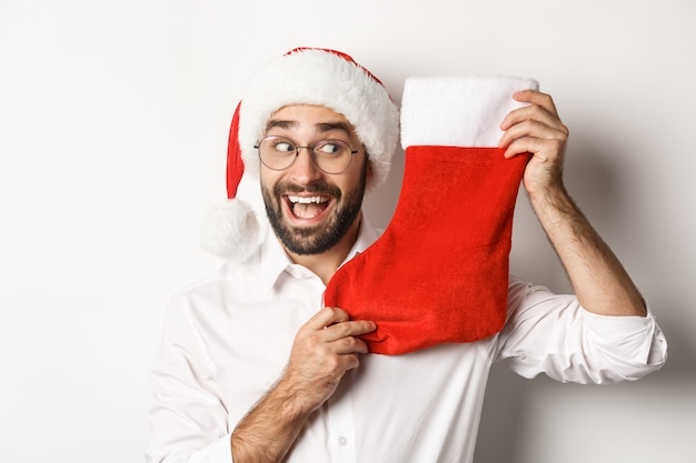 Primer plano de hombre feliz celebrando la Navidad, recibir regalos en calcetines de Navidad y mirando emocionado, con gorro de Papá Noel y gafas, fondo blanco.