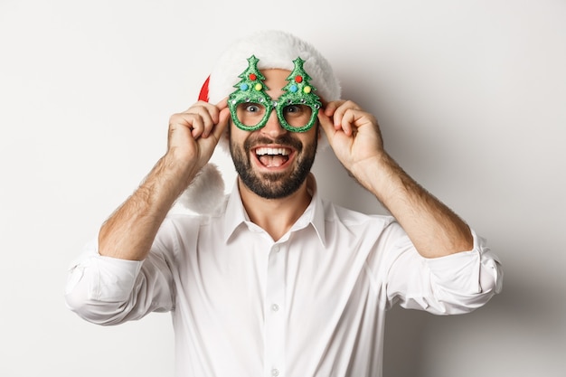 Primer plano de hombre feliz celebrando las vacaciones de Navidad, con gafas de fiesta y gorro de Papá Noel, disfrutando de año nuevo, fondo blanco.