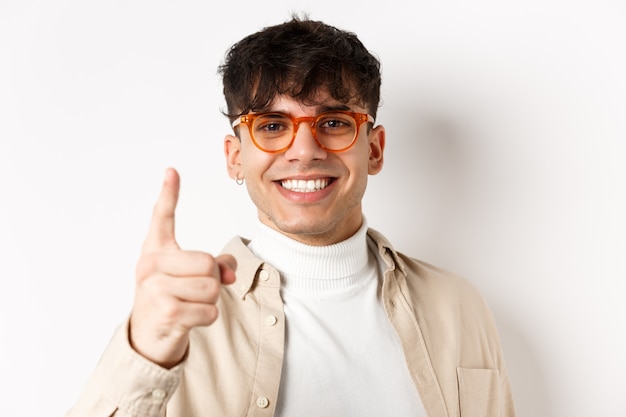 Primer plano de un hombre guapo afortunado con gafas, sonriendo y señalando con el dedo, alabando algo bueno, de pie en la pared blanca.