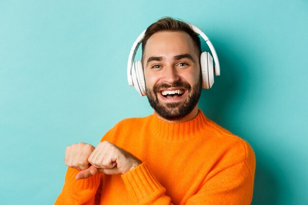 Primer plano de hombre guapo moderno escuchando música en auriculares, de pie en suéter naranja sobre fondo turquesa.
