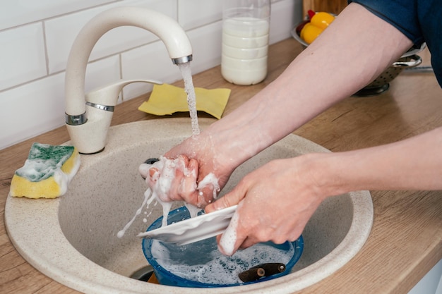 Primer plano de un hombre irreconocible con camisa azul lavando un plato redondo blanco en el fregadero de la cocina de mármol beige bajo el agua corriente del grifo de pie en la cocina moderna blanca Concepto de limpieza de tareas domésticas