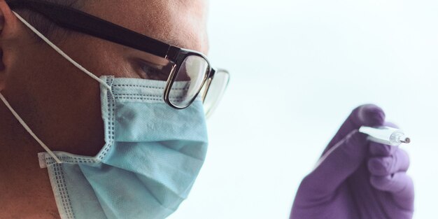 Foto primer plano de hombre joven con gafas, máscara médica y guantes desechables mirando el termómetro.