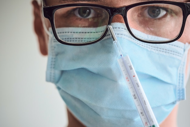 Foto primer plano de hombre joven con gafas, máscara médica y guantes desechables con termómetro.