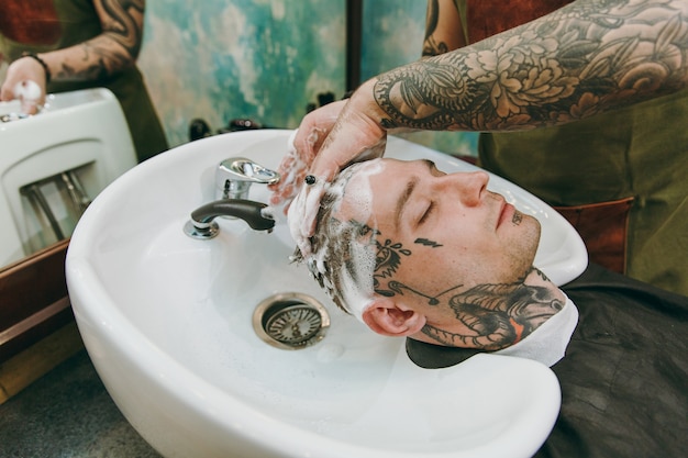 Primer plano del hombre que consigue un corte de pelo de moda en la peluquería. El estilista masculino en tatuajes al servicio del cliente, lavándose la cabeza