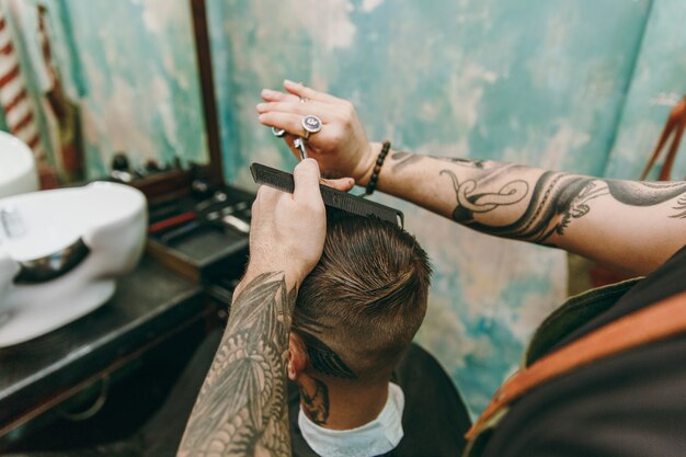 Primer plano del hombre que consigue un corte de pelo de moda en la peluquería. El estilista masculino en tatuajes al servicio del cliente.