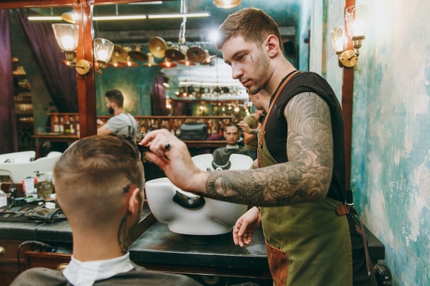 Primer plano del hombre que consigue un corte de pelo de moda en la peluquería. El estilista masculino en tatuajes al servicio del cliente.