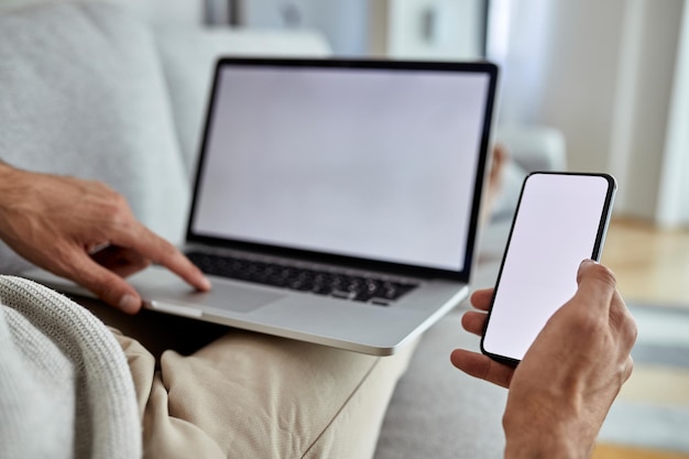 Primer plano de un hombre que usa un teléfono móvil mientras navega por la red en una computadora portátil en casa El foco está en la pantalla del dispositivo en blanco Copiar espacio