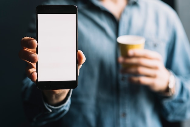 Primer plano de un hombre sosteniendo una taza de café que muestra un teléfono móvil con pantalla blanca