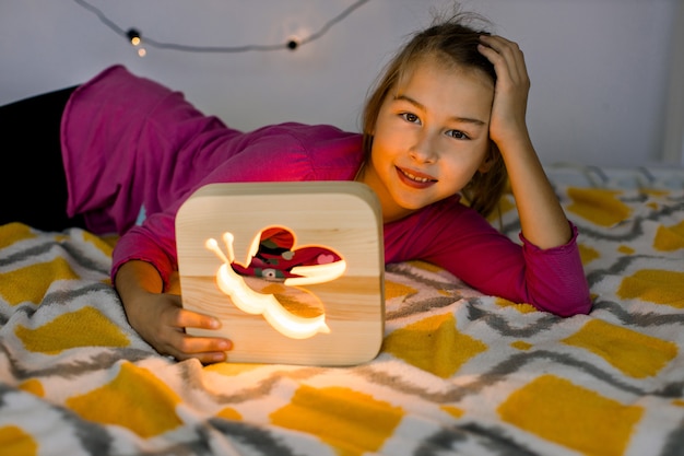 Primer plano horizontal de feliz sonriente niña de 10 años, acostada en una cama acogedora, sosteniendo una hermosa lámpara de noche de madera con imagen de abeja.