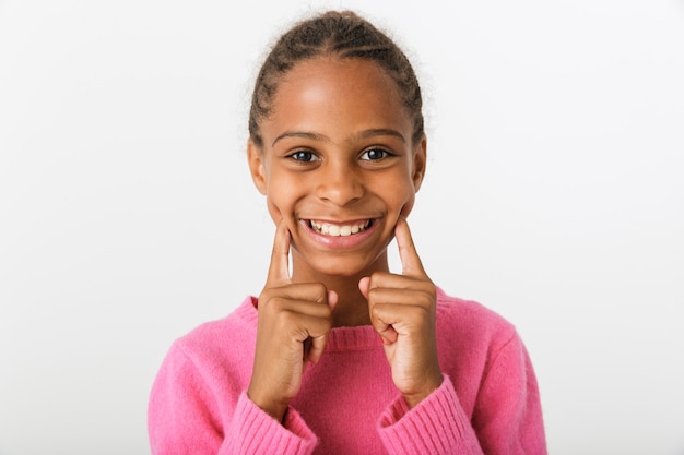 Primer plano de la imagen de la amable niña afroamericana sonriendo y señalando con el dedo a sus mejillas aisladas sobre la pared blanca