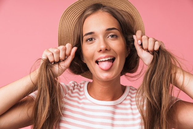 Primer plano de la imagen de una joven linda mujer con sombrero de paja burlándose con su cabello y sacando la lengua aislada sobre pared rosa