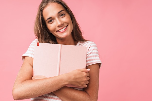 Primer plano de la imagen de una joven mujer bonita sonriendo mientras abraza su diario aislado sobre la pared rosa